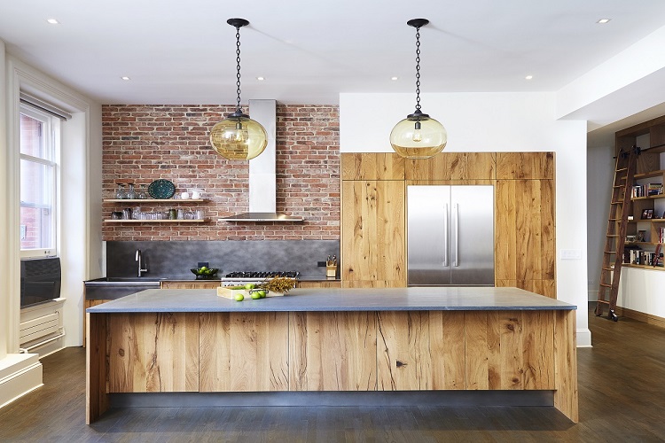 Industrial-modern kitchen with reclaimed wood cabinetry and island, exposed brick wall, stainless steel refrigerator and range, concrete-look countertop, open shelving, and glass pendant lights in a bright loft-style space.