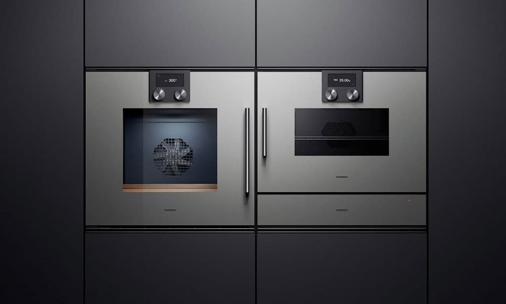 Built-in luxury wall ovens with brushed metal fronts, digital controls, and sleek integrated handles set into dark modern cabinetry.