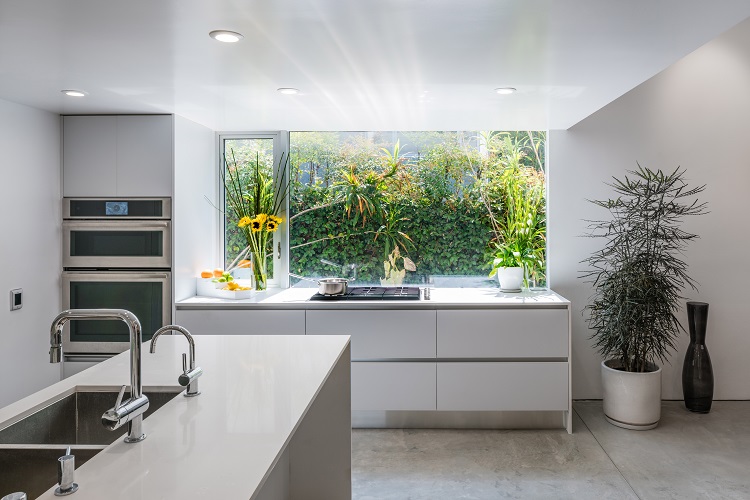 Minimalist modern kitchen with sleek white cabinetry, integrated appliances, waterfall island with built-in sink, large window overlooking greenery, and clean contemporary European kitchen design. by Aster Cucine