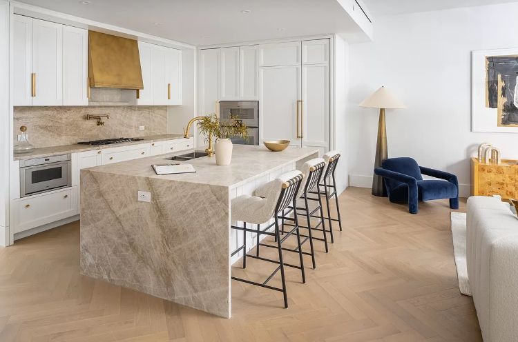 Transitional white kitchen in NYC with custom cabinetry, waterfall stone island, brass hardware, and built-in appliances.